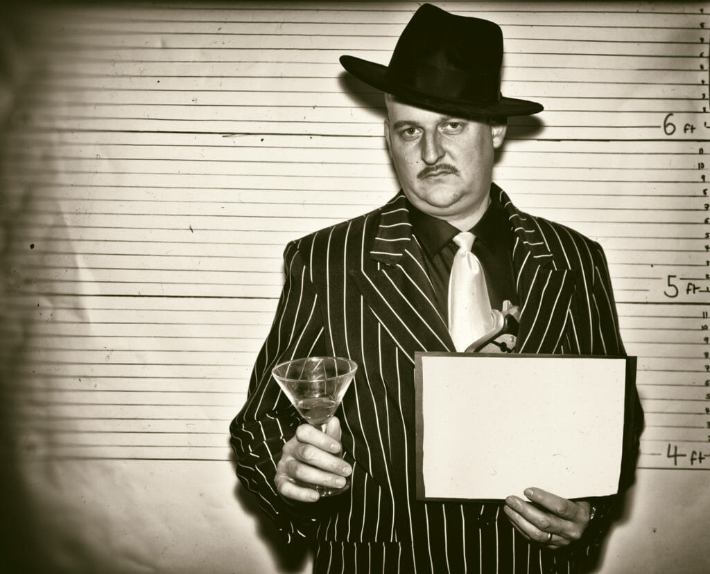 man dressed as old time gangster holding a glass during mugshot
